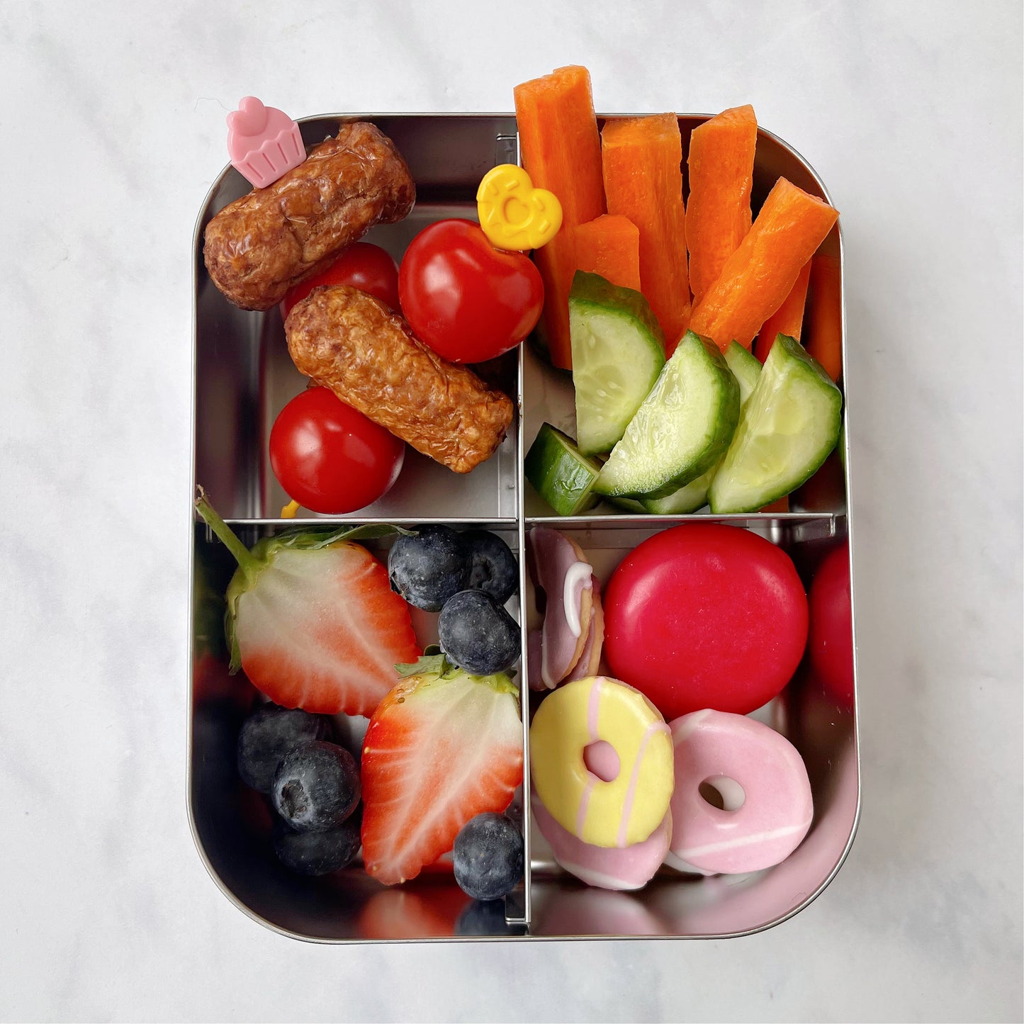Cocktail sausages and cherry tomatoes on a pink and a yellow food skewer in a stainless steel sectioned lunch box that also contains chopped fruit and veg as well as some mini party rings.