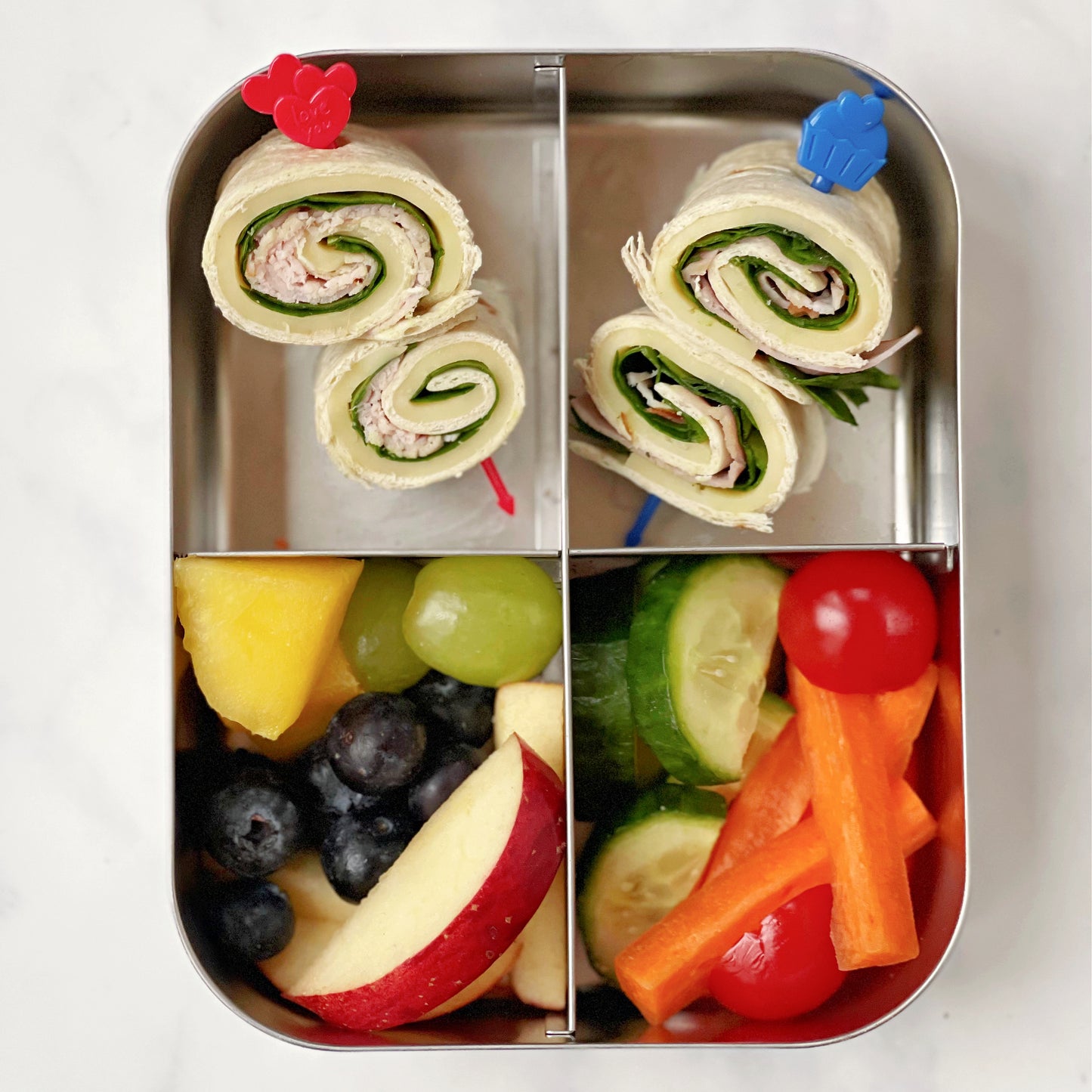 a four section stainless steel lunchbox with fresh chopped fruit and veggies and wraps held together with Pick Sticks food skewers for kids.