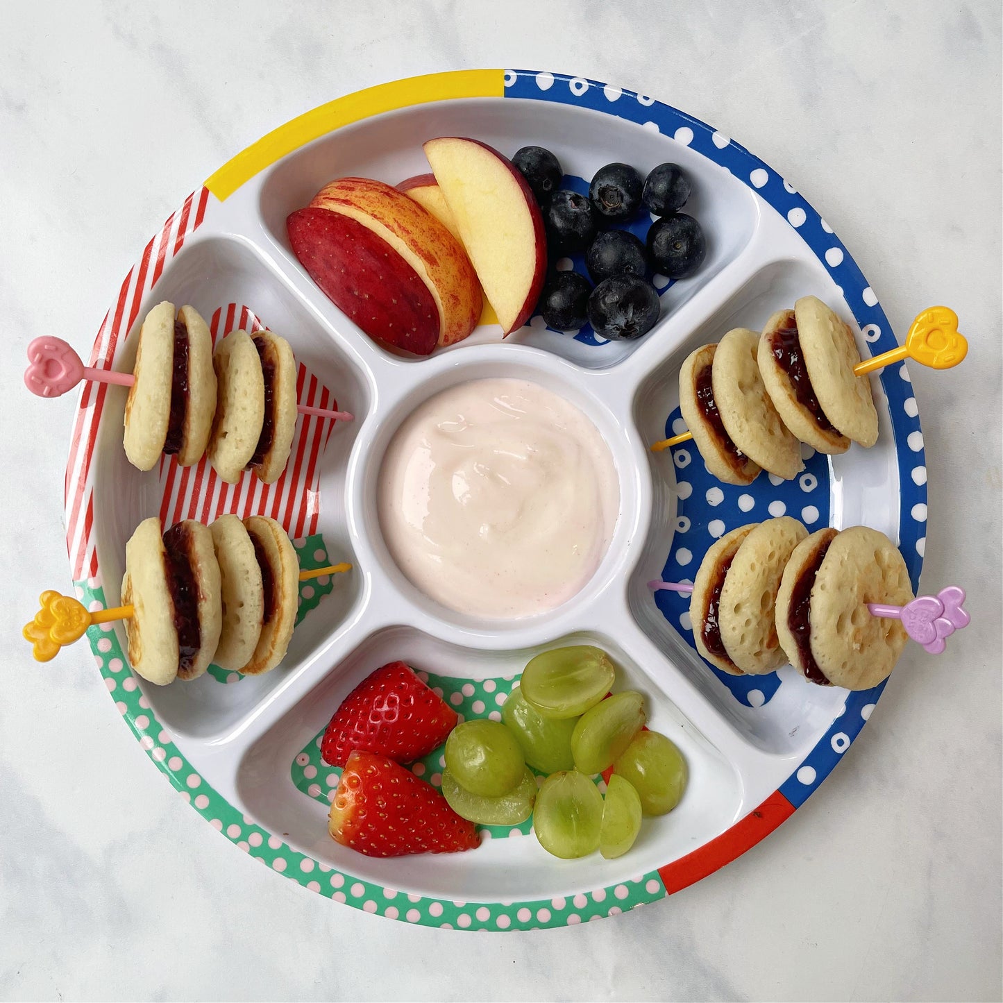 Mini pancakes on food skewers in a colourful five compartment plate.