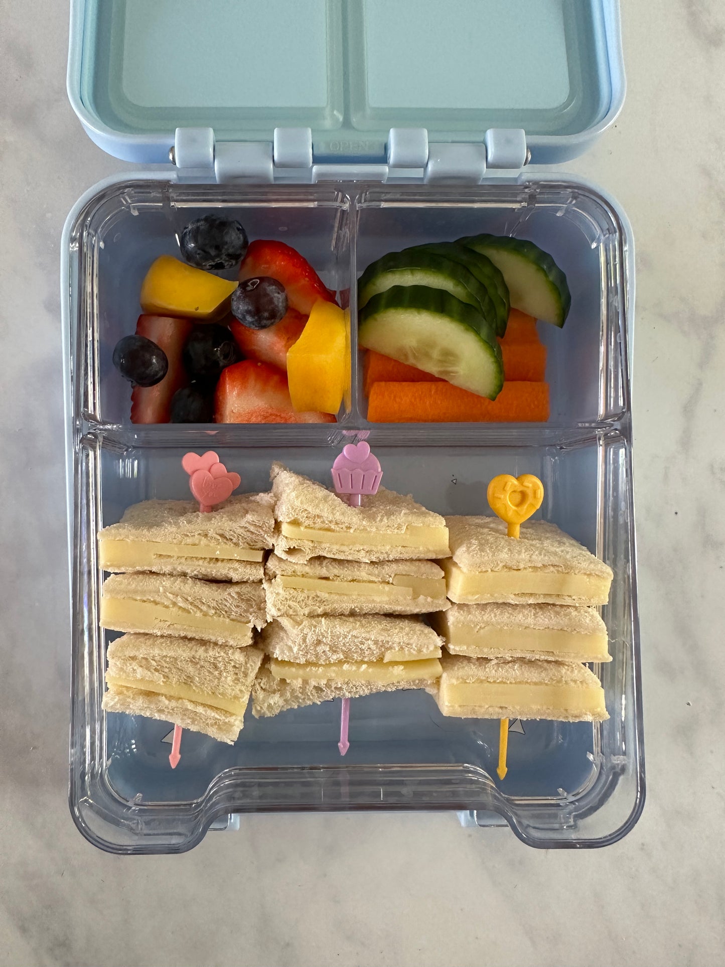 A square compartment lunchbox showing how Pick Sticks Pastels have been used to make sandwich skewers. Also with fresh chopped fruit and veggies in the other sections of the lunchbox.