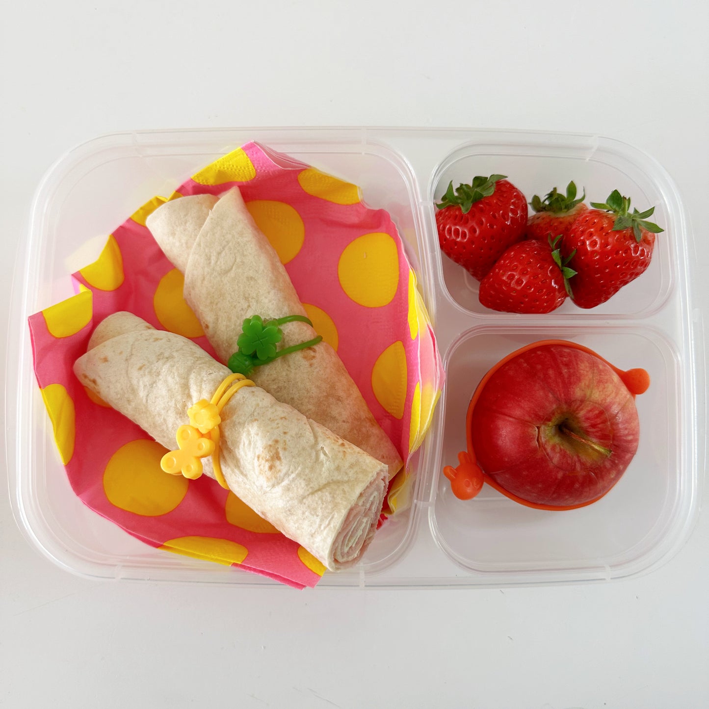 A filled white wrap cut into half and secured with one green and one yellow wrap band in a clear divided lunch box with an apple and strawberries.