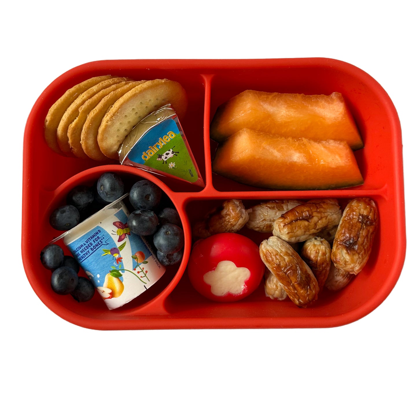 a red silicone lunchbox for kids with sections filled with cocktail sausages, a mini Babybel cheese with a flower shape cut out of the red wax covering, a Dairylea cheese triangle, mini cheese crackers, sliced cantaloupe melon, blueberries and a small kids yogurt.