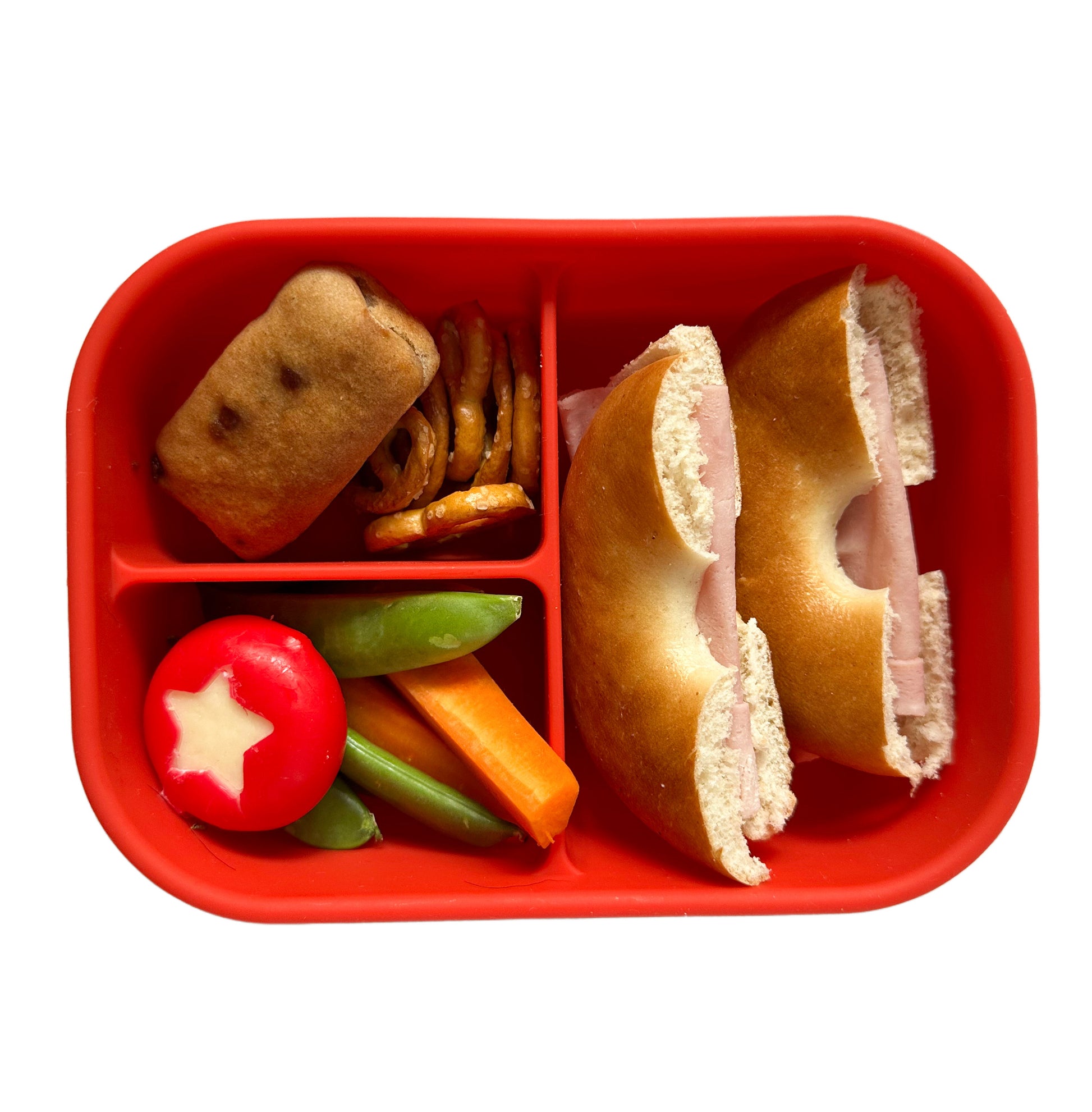 a red sectioned lunchbox containing a ham bagel, pretzels, a mini cake bar, sugar snap peas, carrot batons and a mini Babybel cheese with a star shape cut out of the red wax covering.