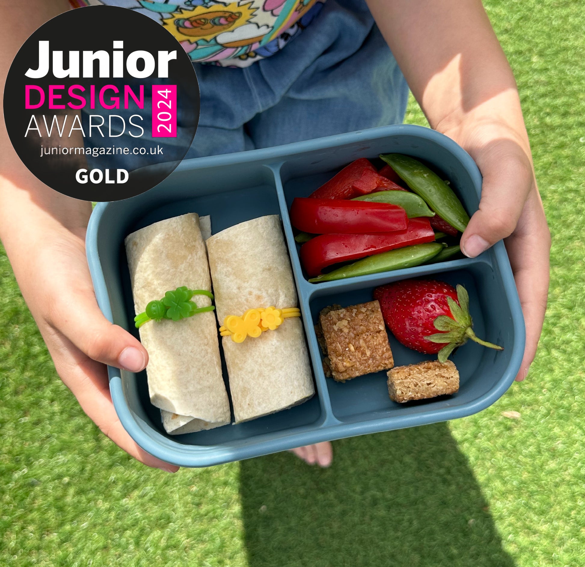 a child holding a blue 3 compartment lunchbox filled with a wrap held together with a wrap band, chopped vegetables, a strawberry and two small squares of flapjack. Also bearing the Junior Design Awards Gold 2024 winner badge.