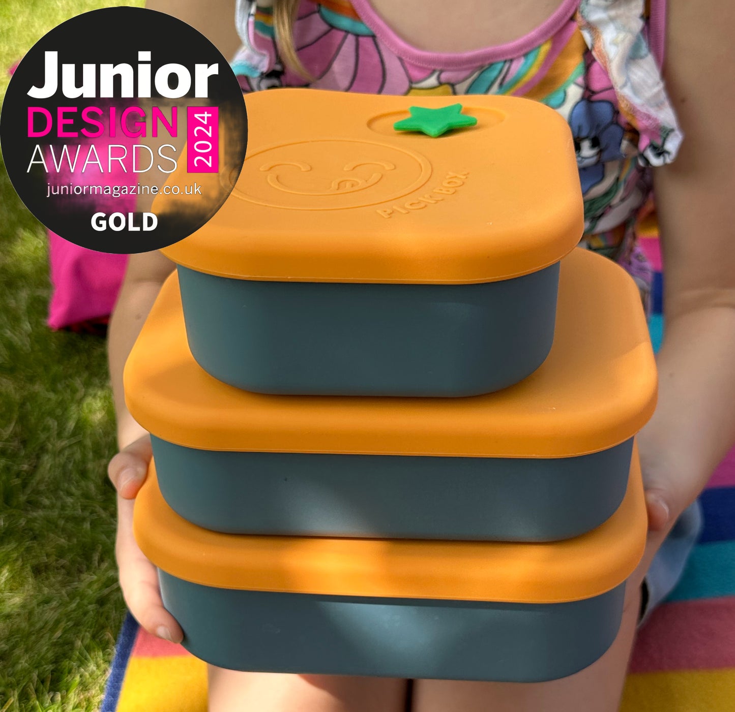 A child on a picnic blanket holding 3 blue and yellow silicone lunchboxes in three different sizes and showing the Junior Design Awards 2024 Gold Winner Badge.