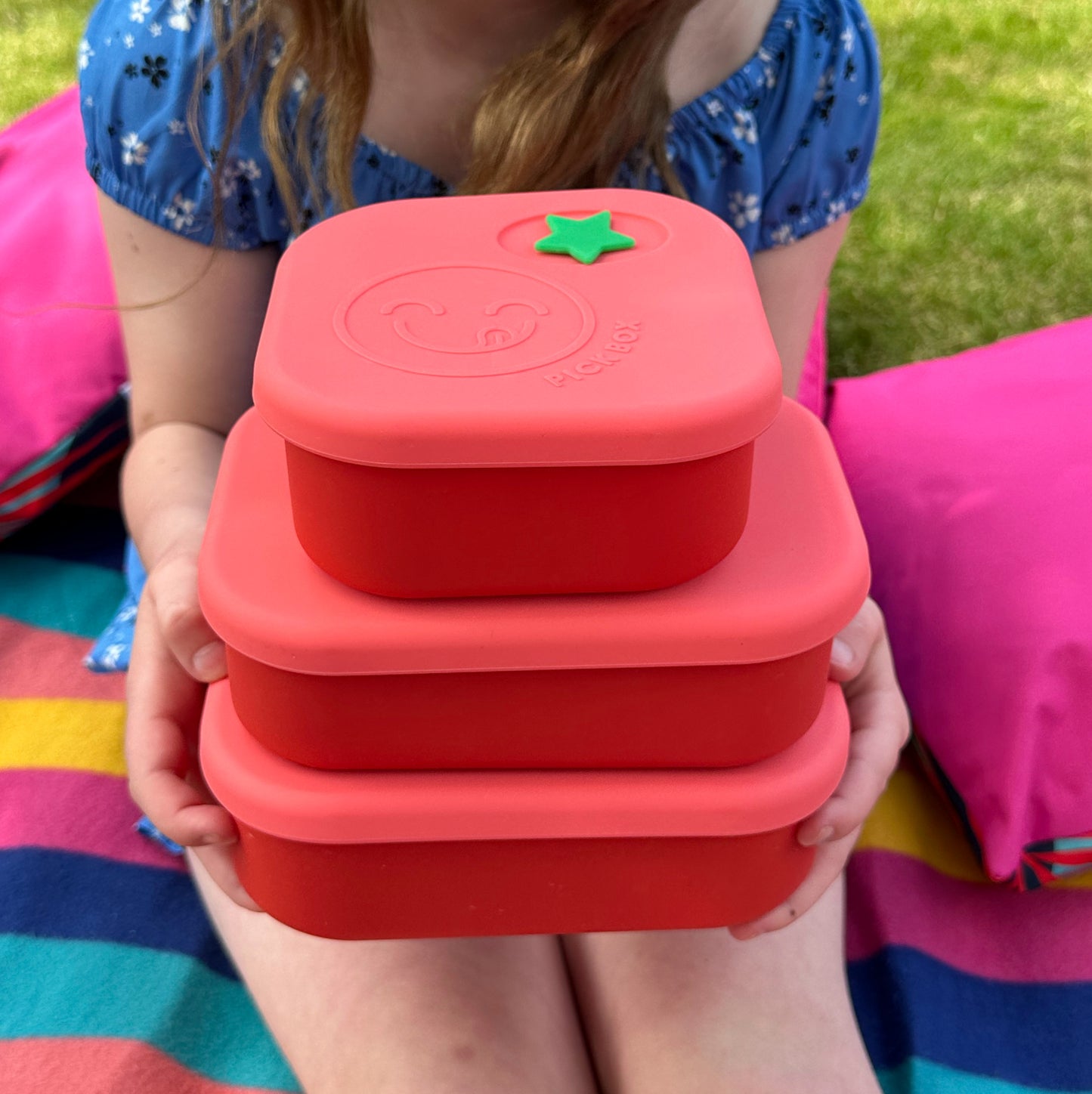 A child on a picnic blanket holding 3 pink and red silicone lunchboxes in three different sizes.