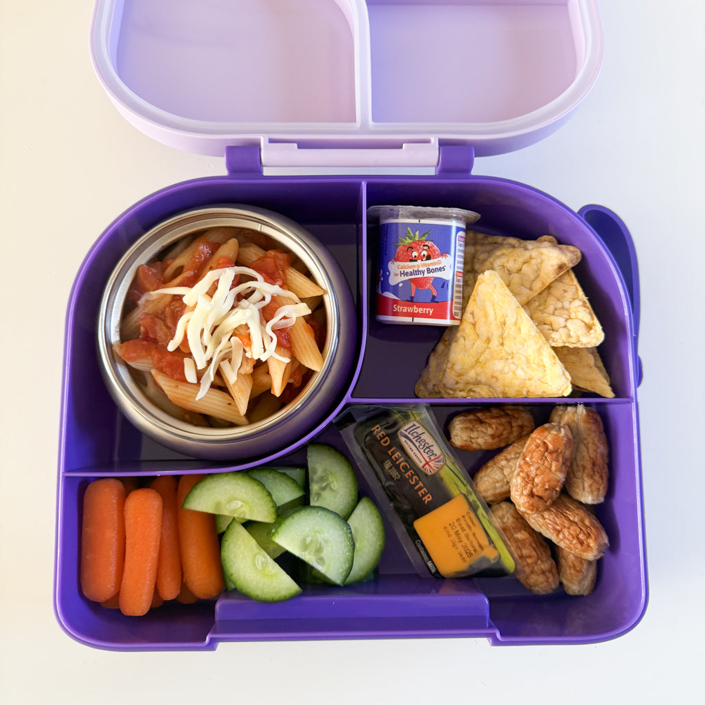 A purple divided lunchbox with a stainless steel food thermos inside. Packed with cooked pasta and cheese, a Petit Filous yogurt, cocktail sausages, mini Red Leicester cheese, chopped carrot and cucumber and some tortilla chips.