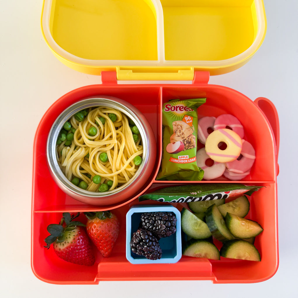 A yellow and orange divided lunchbox with a stainless steel food thermos inside. Packed with noodles, Soreen bar, fresh fruit and veggies, party rings and a small pepperami.