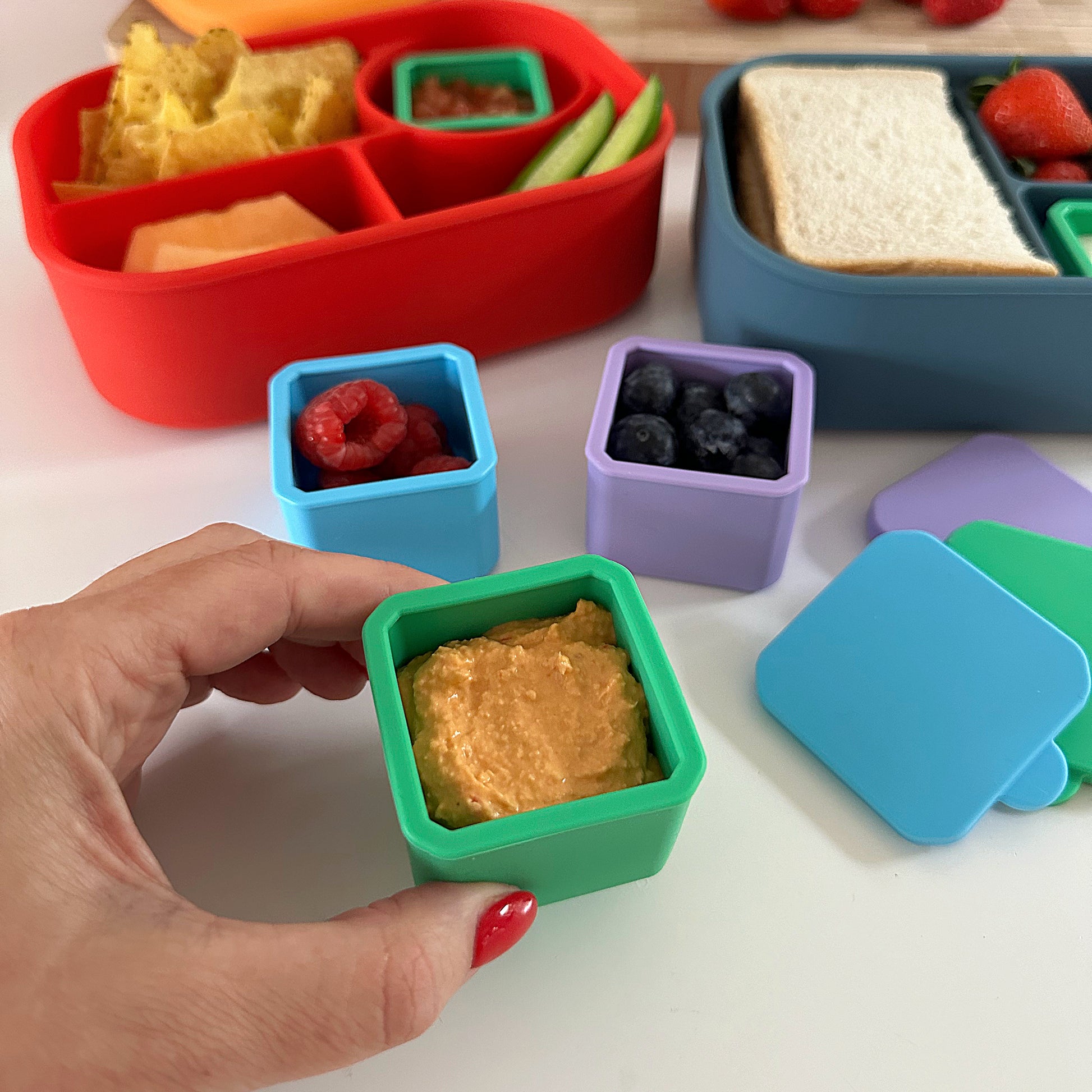 3 small silicone food containers with lids containing hummus, raspberries and blueberries next to brightly coloured silicone lunchboxes from Pick Plates.