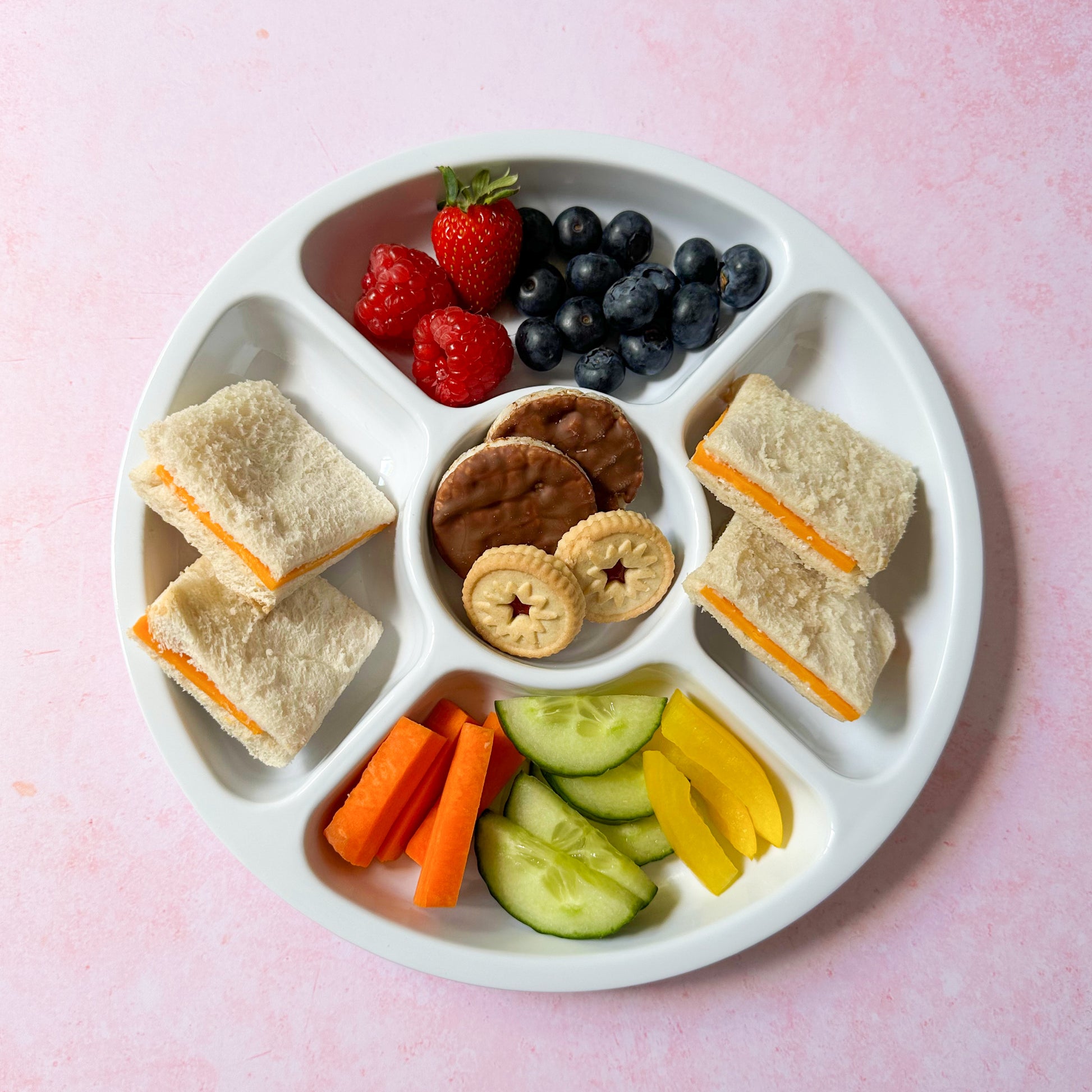white compartment plate for kids filled with a cheese sandwich, chopped fruit and veg, chocolate covered rice cakes and mini jammy dodgers.