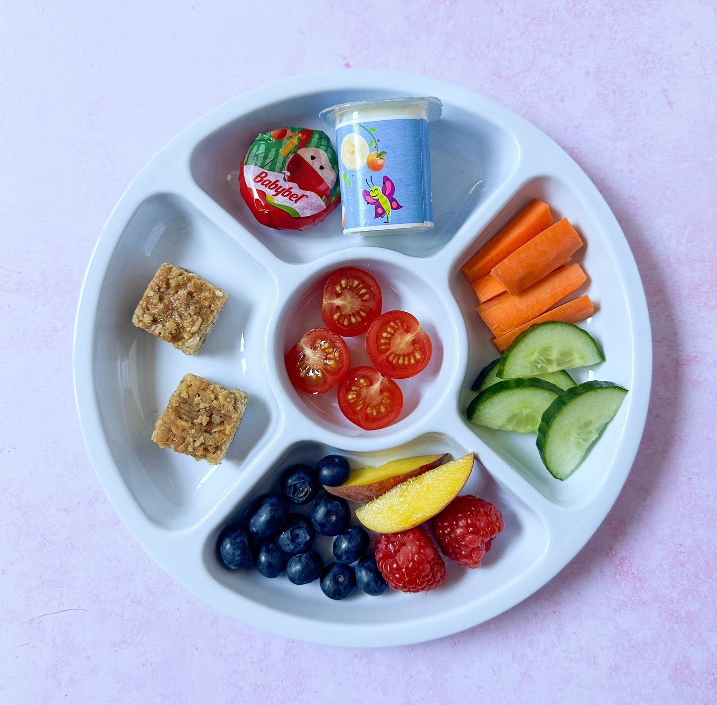 white sectioned plate for kids filled with chopped fruit and veg, a mini Babybel cheese, small yogurt pot and two small squares of flapjack.