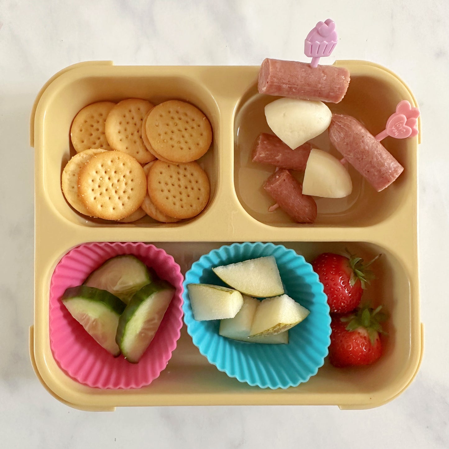 A pink and a purple kids food skewer with two slices of Pepperami and a quarter of Mini Babybel cheese on each. In a yellow compartment snack box also containing chopped fruit and veg and mini cheedars.