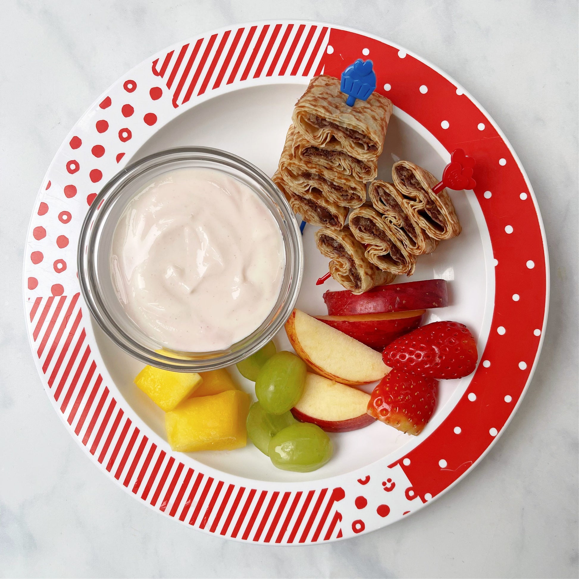 A red and blue Pick Stick with a chocolate filled crepe on each skewer, served on a classic range children's plate with a serving of yogurt and fresh fruit. 