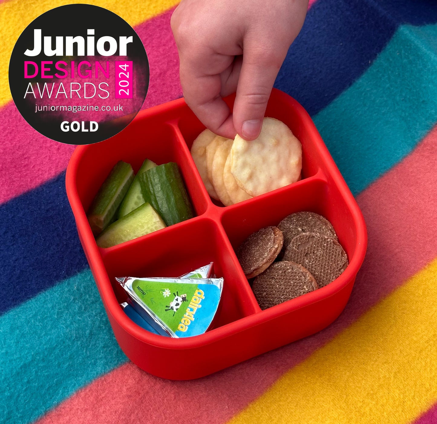 a four compartment red snack box filled with rice crackers, Dairylea cheese, mini chocolate biscuits and cucumber batons with the image also showing the showing the Junior Design Awards 2024 Gold Winner Badge.