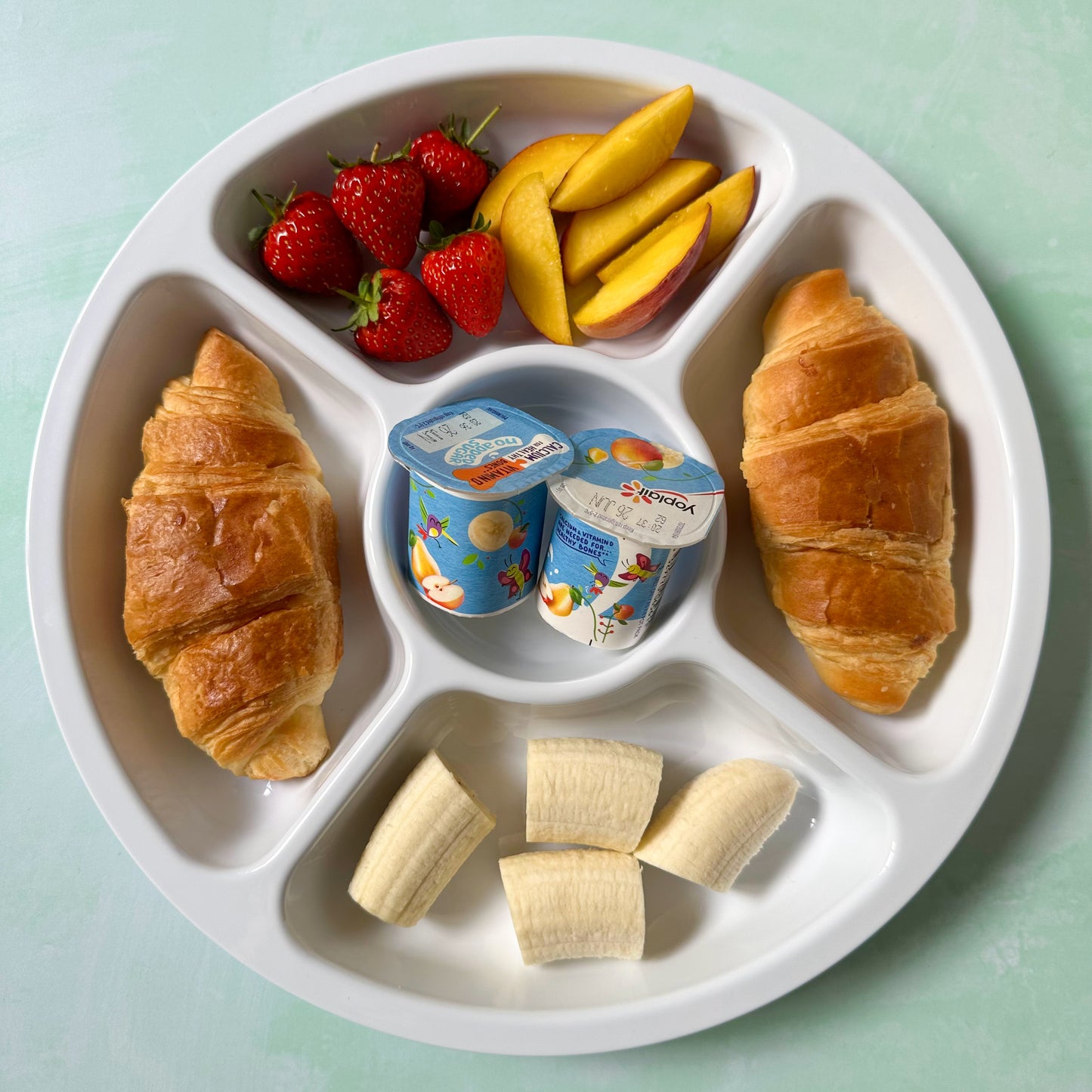 A large 5 section compartment plate for picky eaters filled with croissants, chopped banana, peach slices, strawberries and two small yogurt pots.