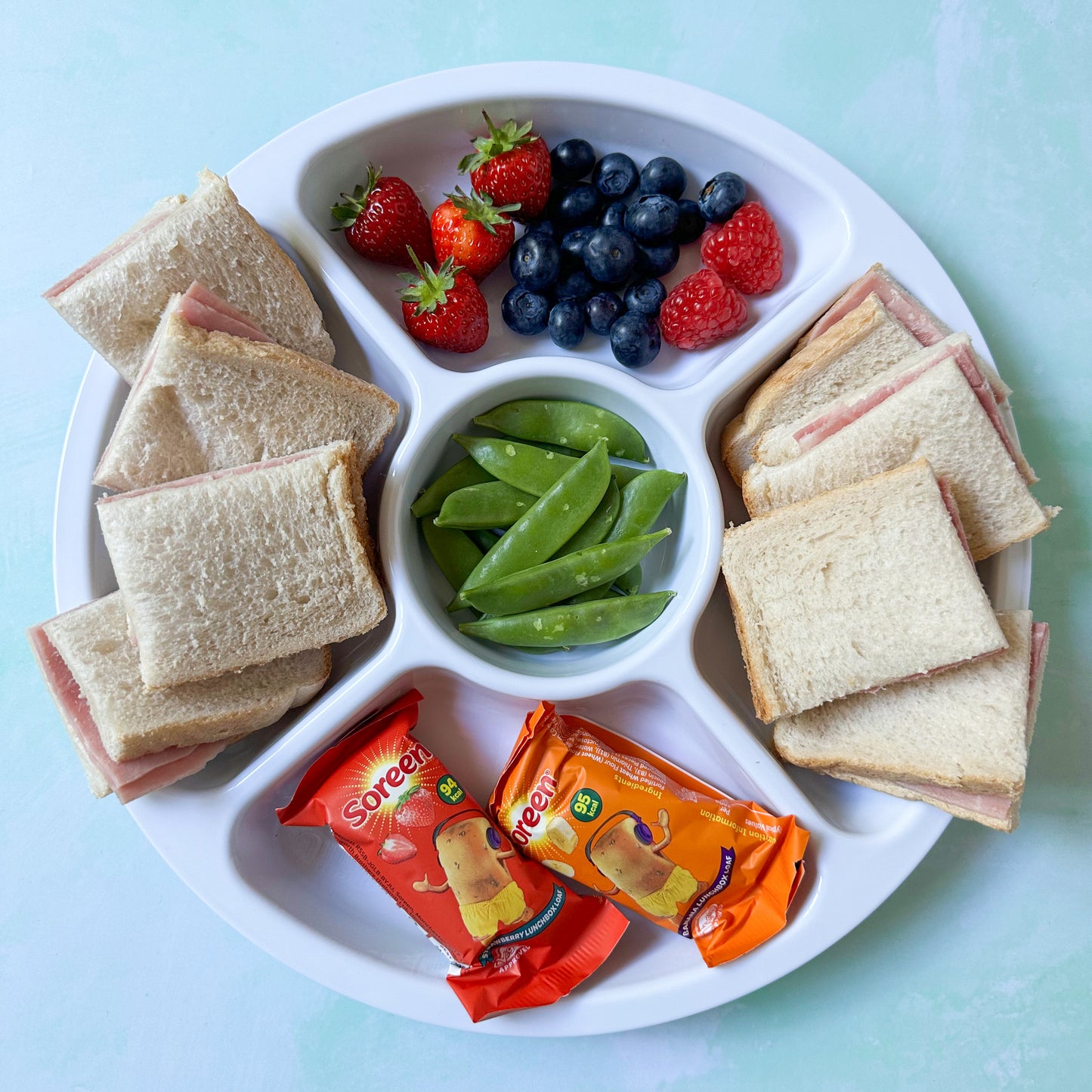 Large white sectioned plate for fussy eaters filled with ham sandwiches, sugar snap peas, Soreen Bars, fresh strawberries and blueberries.