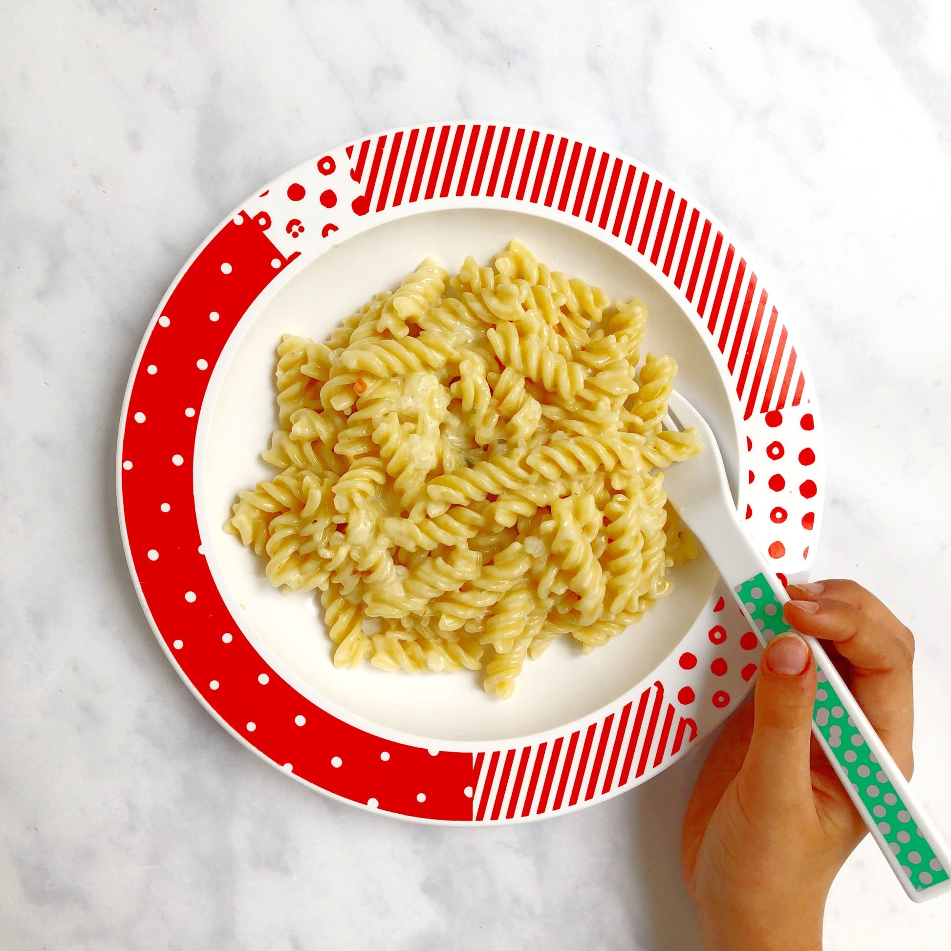 red and white patterned children's plate with a portion of pasta and a child holding a fork in the pasta