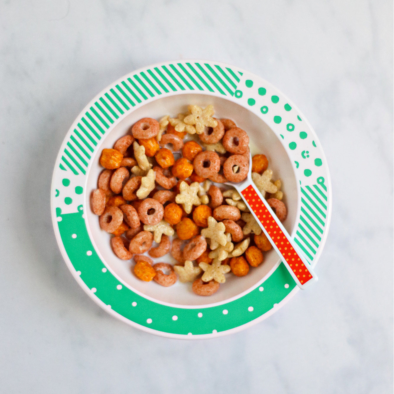 white and green patterned melamine bowl containing cereal and milk with a brightly coloured kids spoon in it.