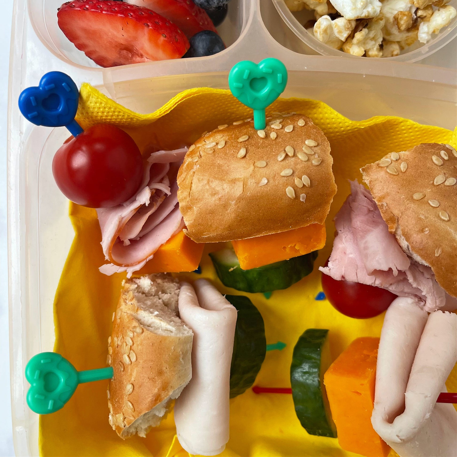 Four Pick Sticks bering used for bagel slices, cheese, ham, cucumber and tomato in a plastic lunchbox.