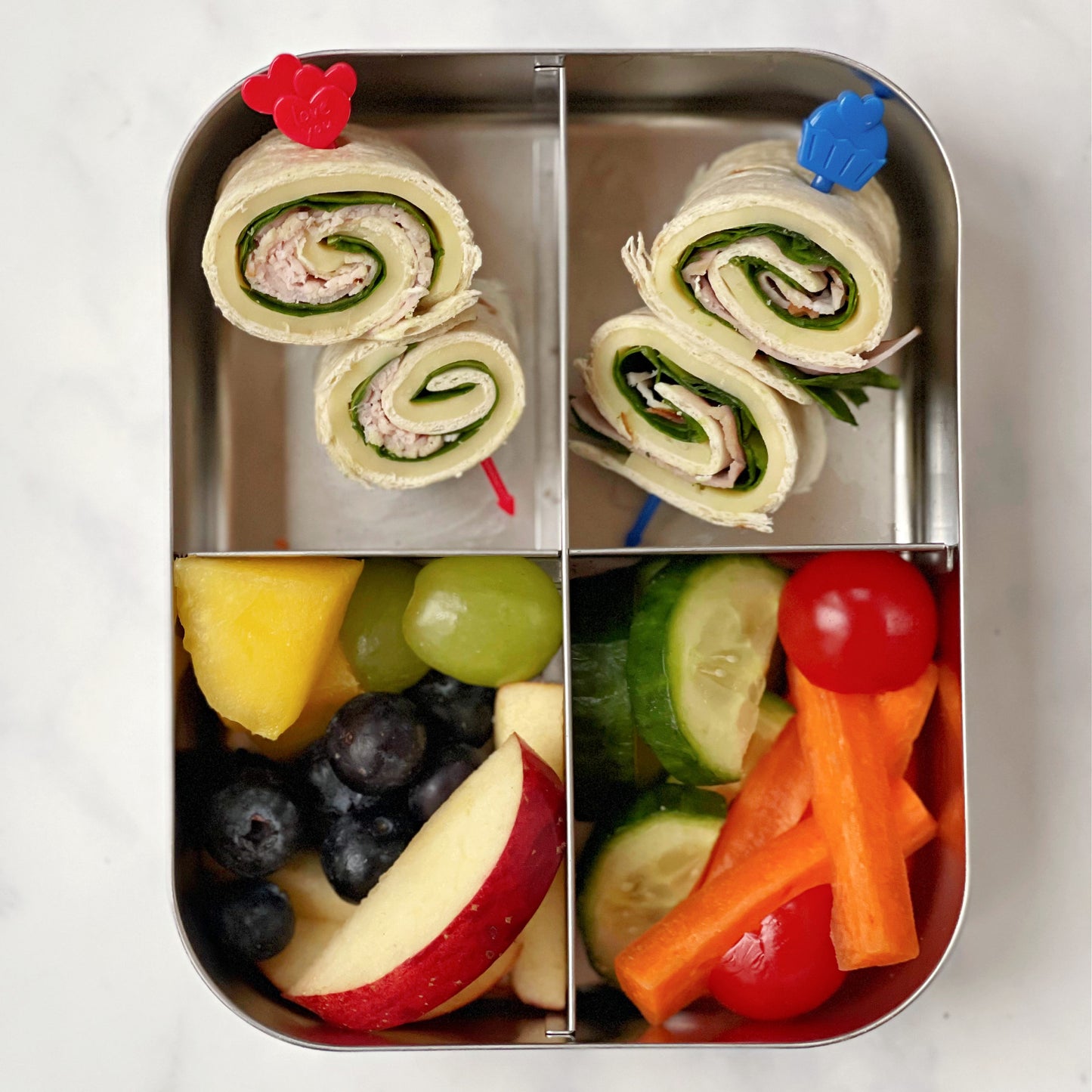 A red and blue Pick Stick with a filled wrap on each skewer in a four comparment metal lunchbox and a serving of fresh fruit and vegetables.
